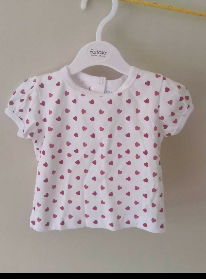 White baby shirt with pink heart pattern on a hanger against a beige wall.