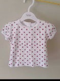 White baby shirt with pink heart pattern on a hanger against a beige wall.