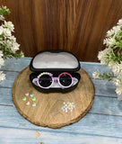Sunglasses with decorative elements in a black case on a wooden surface with flowers around.