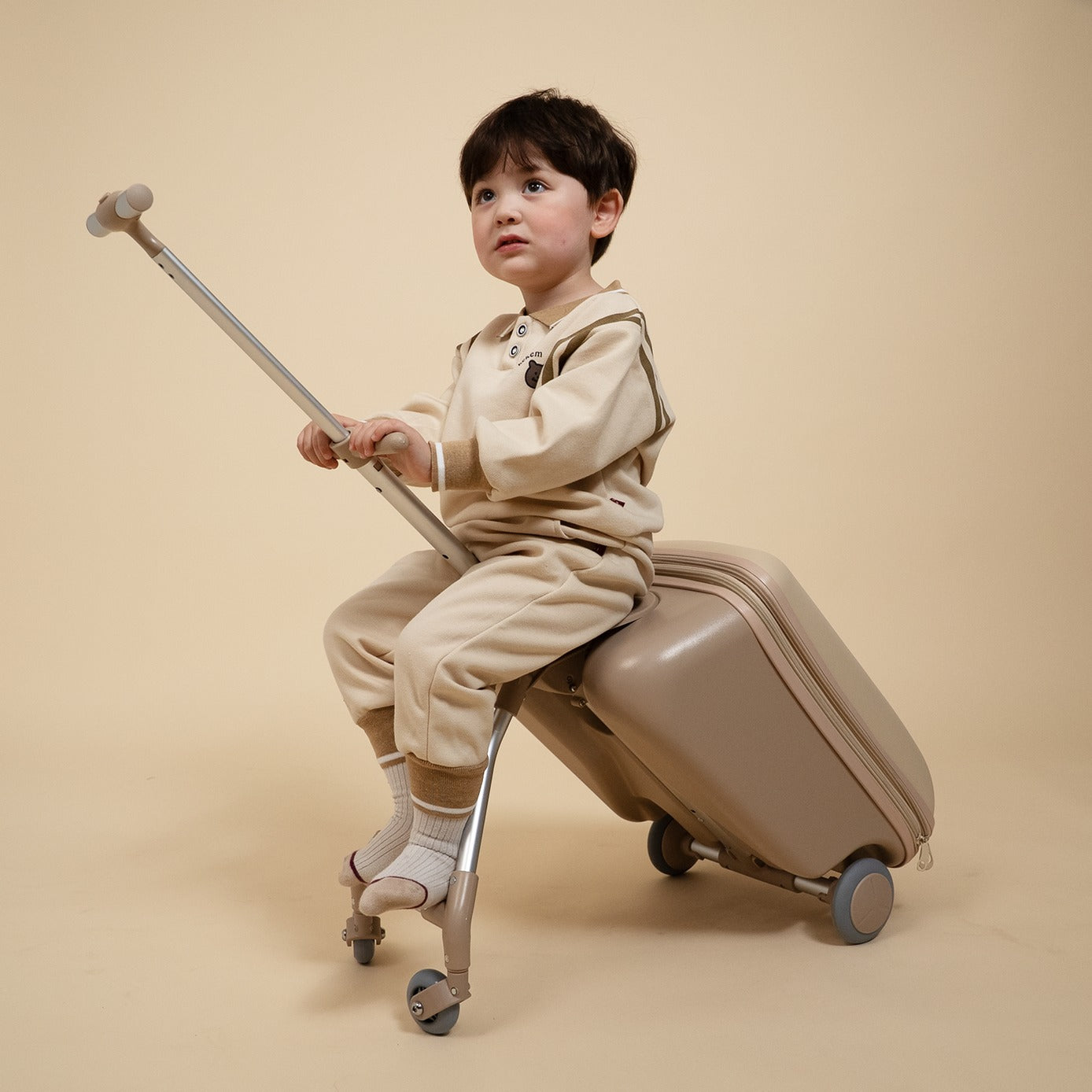 Child sitting on a suitcase with a rolling toy, both in beige, against a beige background
