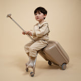 Child sitting on a suitcase with a rolling toy, both in beige, against a beige background