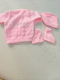 Pink baby sweater, hat, and booties set on a light gray surface