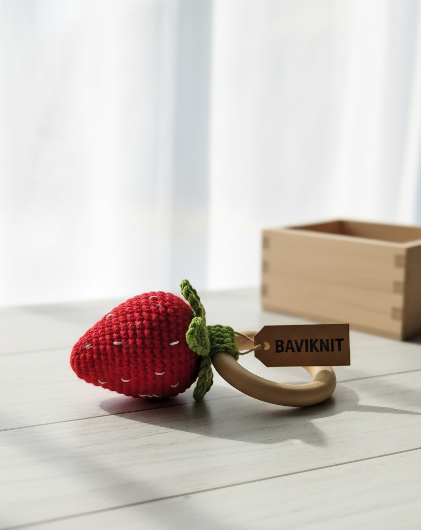 Red strawberry-shaped teething ring with a wooden tag labeled 'BAVIKNIT' on a light wooden surface.