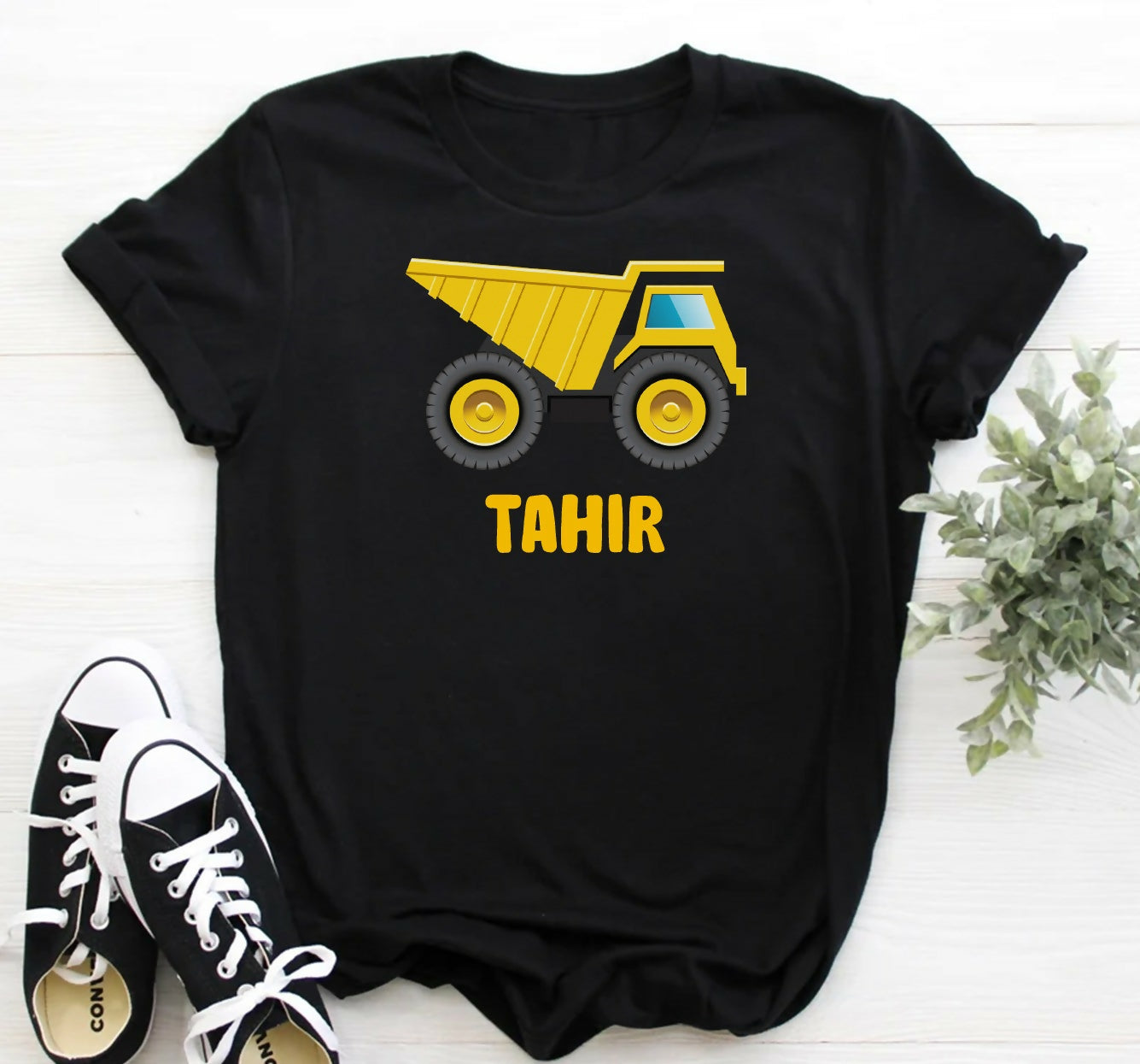 Black t-shirt with yellow dump truck graphic and 'Tahir' text, paired with black and white sneakers on a light wooden surface.