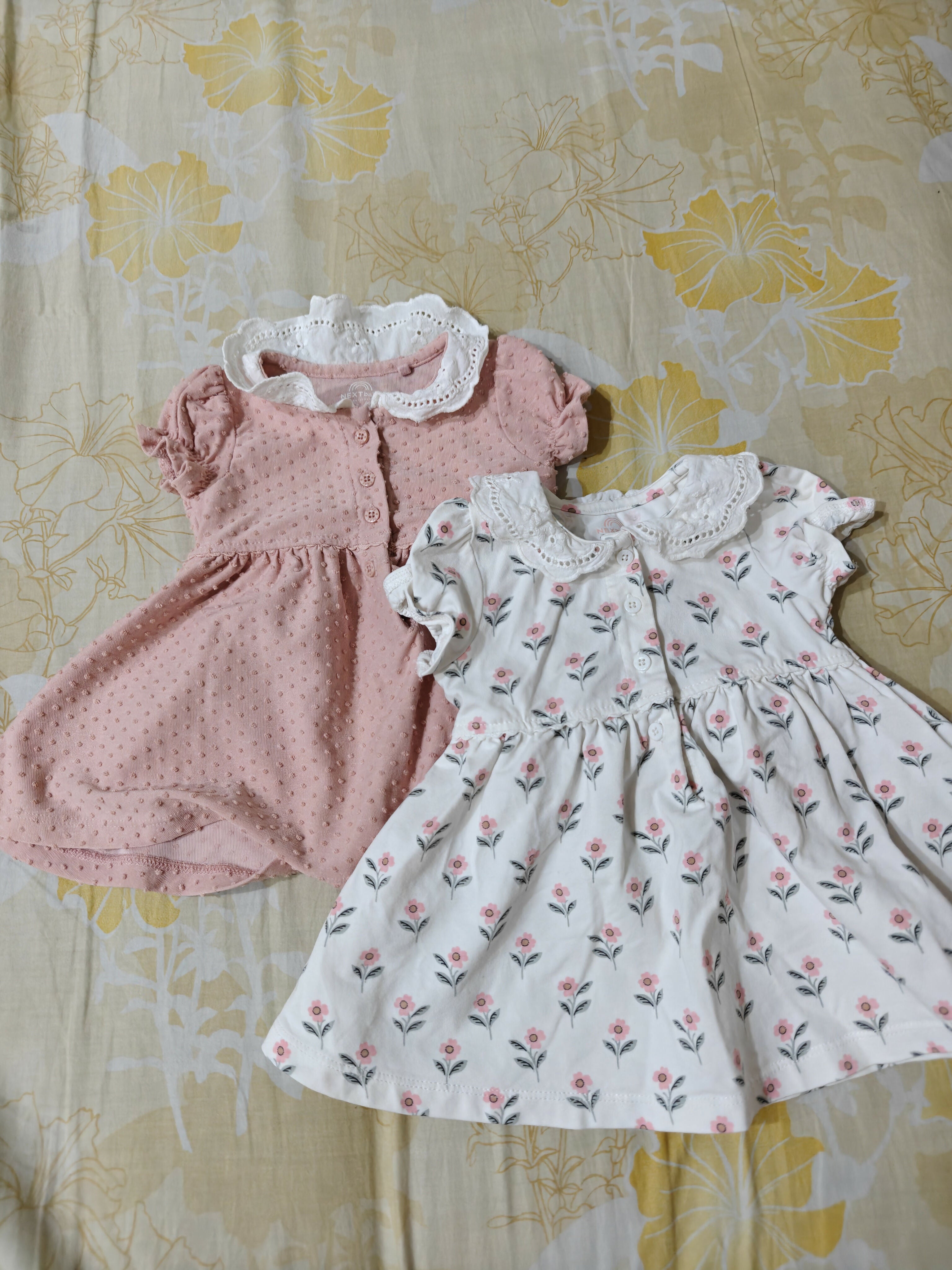 Two children's dresses, one pink and one floral, on a floral-patterned surface.