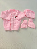 Pink baby cardigan, hat, and booties set on a textured surface