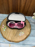 Pink sunglasses with decorative elements in a black case on a wooden surface.