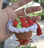 Hand holding a crocheted keychain shaped like a cupcake with red roses, green leaves, and a red bow.