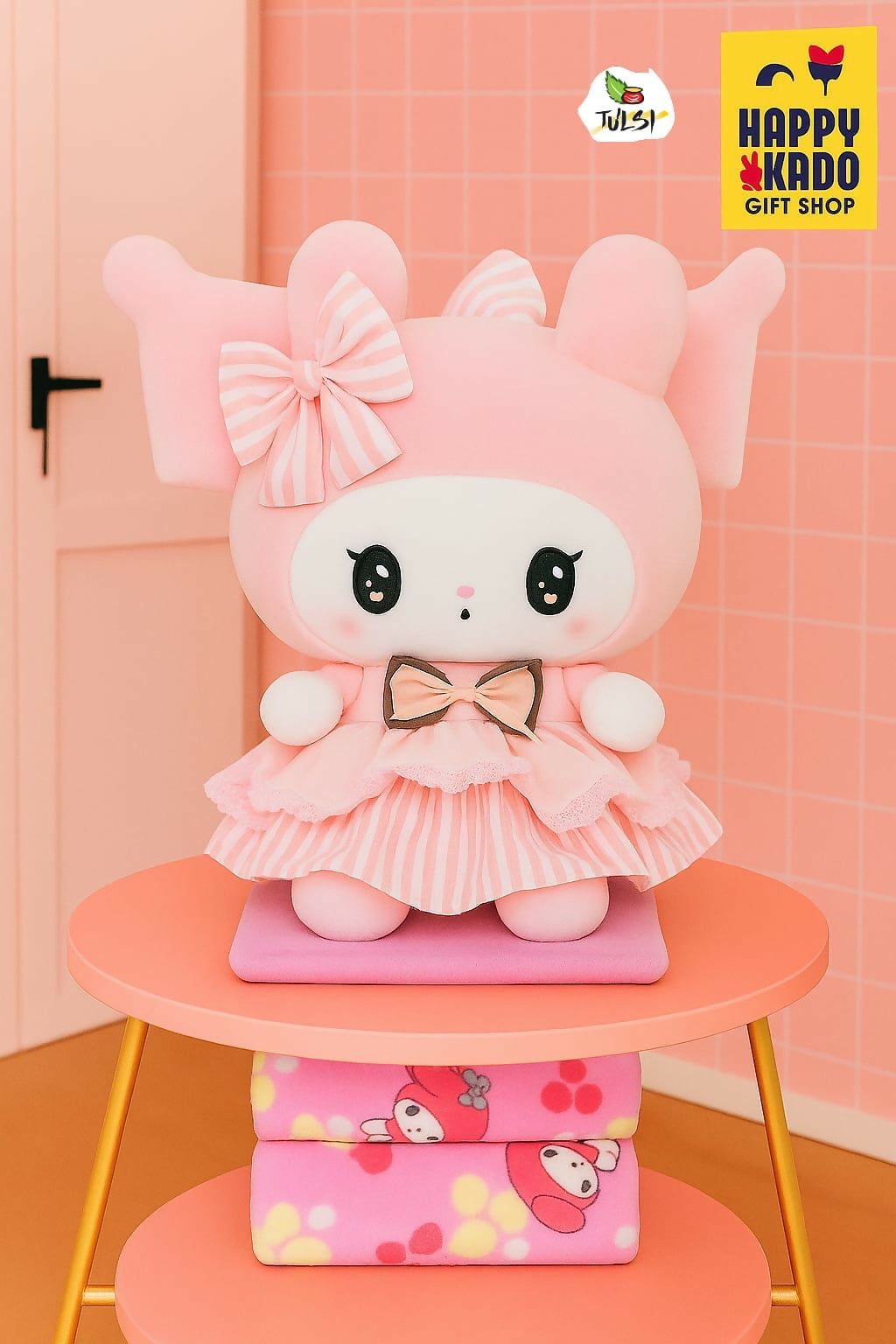 Pink cartoon plush toy with a bow on a pink stool against a pink wall with 'Happy Kado' branding.