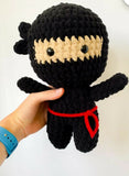 Hand holding a black crochet ninja doll with a red belt against a white background