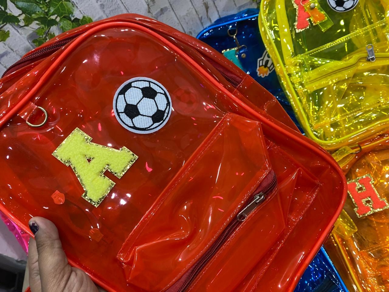 Red transparent backpack with soccer ball design and letter 'H' held by a hand, with other colorful backpacks in the background.