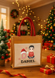Gift bag with 'DANIEL' printed on it, featuring a cartoon design of a person in a Santa hat and a child, set against a festive background with Christmas trees and lights.