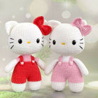 Two crochet Hello Kitty dolls, one in red overalls and the other in pink, against a blurred background.