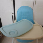 Children's high chair with blue cushion and beige tray against a white wall.