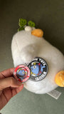 White plush toy with two badges held by a hand on a neutral background