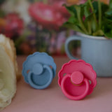 Two colorful pacifiers on a surface with flowers and a cup in the background
