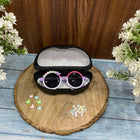 Sunglasses with decorative elements in a black case on a wooden surface with flowers around.