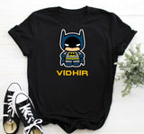 Black t-shirt with a cartoon Batman graphic and 'VIDHIR' text, paired with black and white sneakers on a light wooden surface.