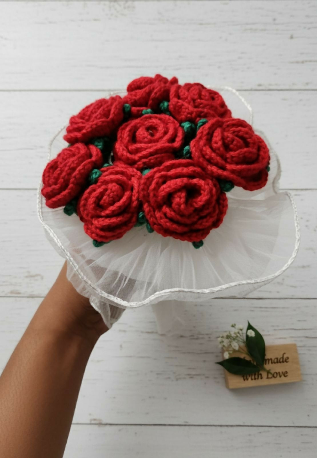 Bouquet of red knitted roses on a white stand with a wooden tag below.
