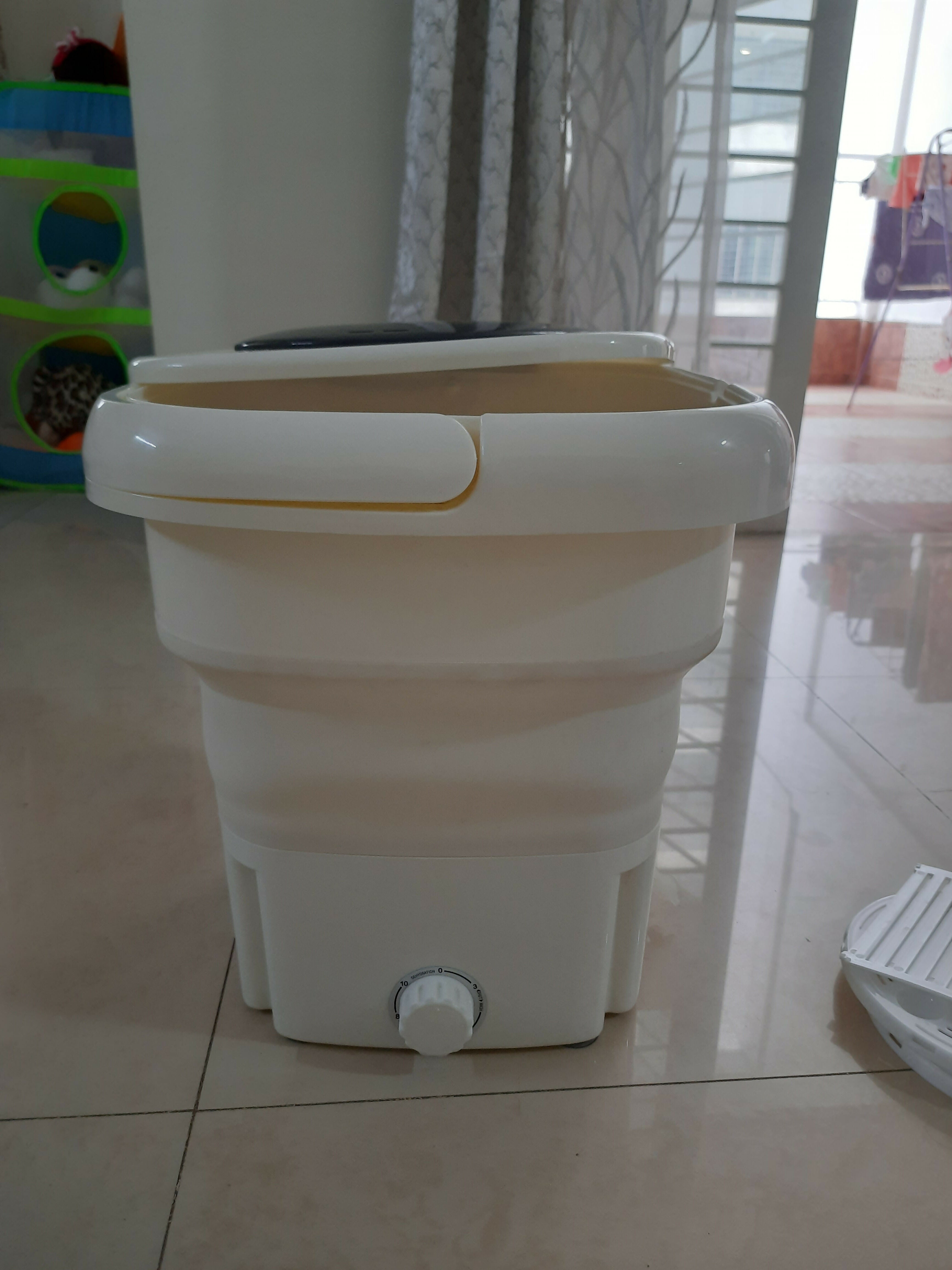 Collapsible washing machine on a tiled floor with curtains in the background
