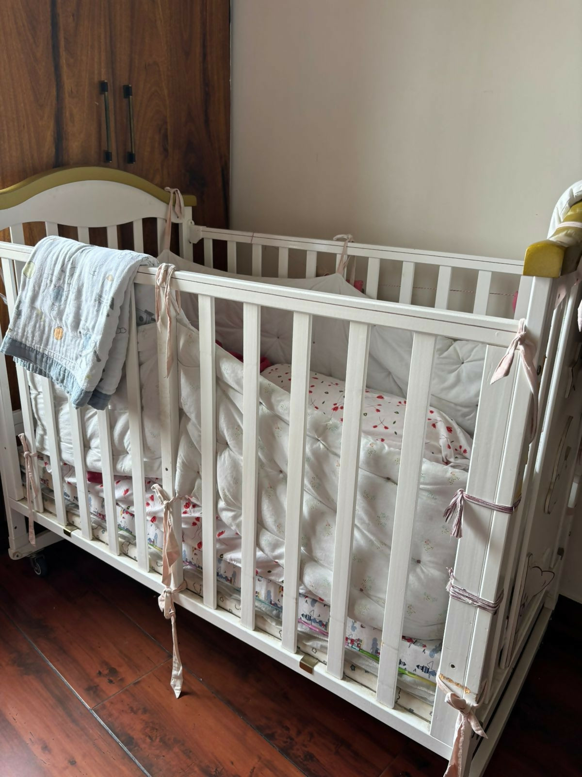 White crib with baby blankets and pillows in a room with wooden floor and white wall.
