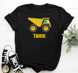 Black t-shirt with yellow dump truck graphic and 'Tahir' text, paired with black and white sneakers on a light wooden surface.