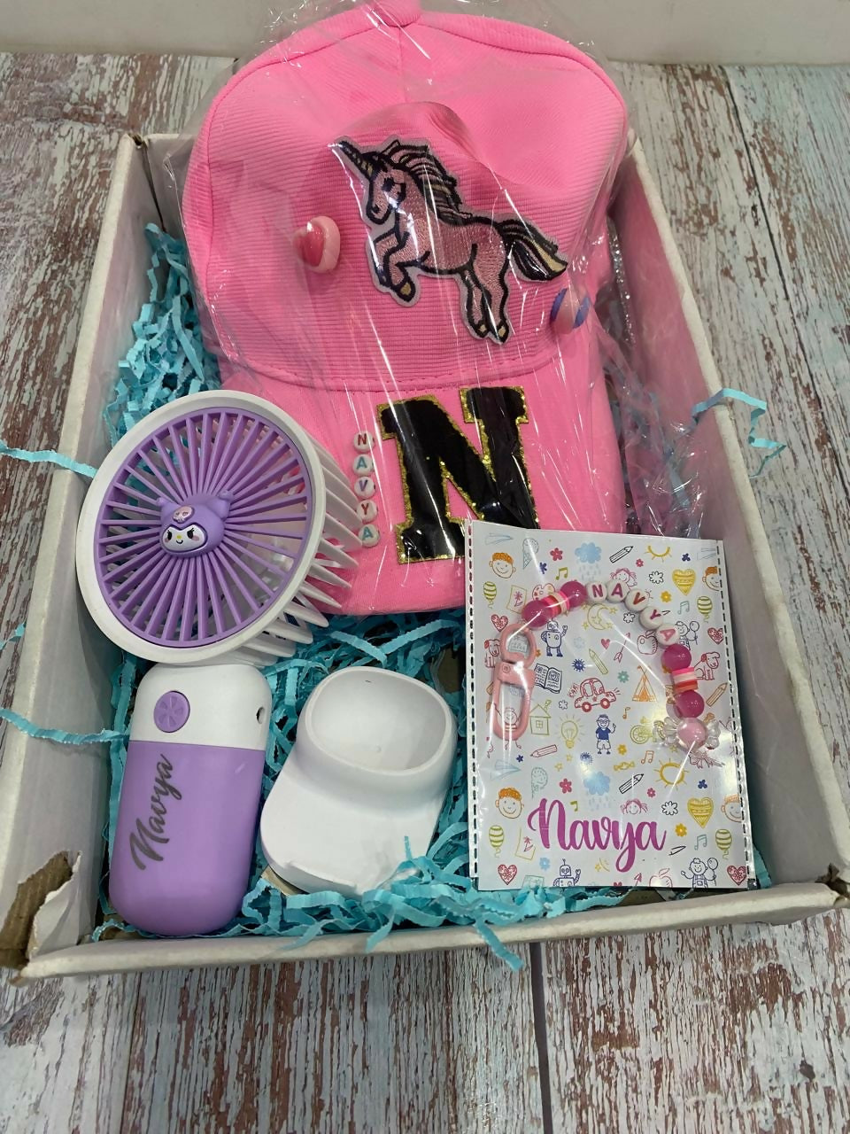 Box containing a pink cap with a horse design, a purple and white toy, a small white container, and a book with a pony design on a wooden surface.