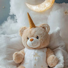 Teddy bear with unicorn horn sitting on a fluffy white surface with a moon and stars in the background.