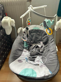 Gray baby bouncer with toys and a white cord on a wooden floor.