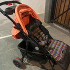 Black stroller with orange cover and colorful striped footmuff on a stone pavement.
