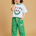 Child wearing a white t-shirt with a smiley face and green pants 