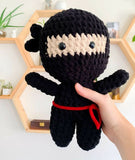 Handmade crochet ninja doll held by a hand with a wooden shelf background