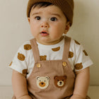 Baby wearing a brown knit hat and bear-themed overalls sitting on a white surface.